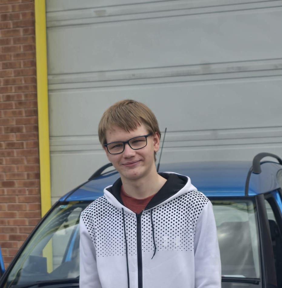 A person with short light/brown hair and glasses stands in front of a blue car and a gray garage door